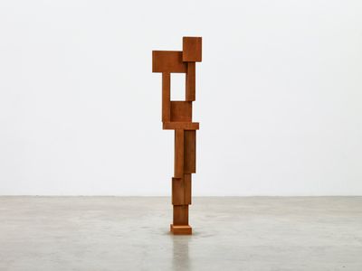 A tall, narrow sculpture in rust-brown is erect in a white gallery space.