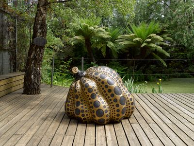 A black and gold pumpkin-shaped sculpture by Yayoi Kusama sits on decking outdoors. There is lush green vegetation behind, including a pair of ferns.