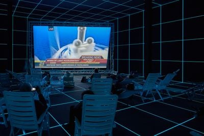 A futuristic looking theatre showing a row of people sitting on lounge seats and facing  big screen showing a single-channel video work by artist Hito Steyerl, entitled Factory of the Sun at the 56th Venice Biennale 