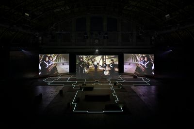 An darken industrial space with three big screens showing a three-channel video work by artist Hito Steyerl at Park Avenue Armory, NYC