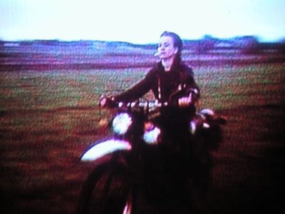 A grainy video still depicting a person riding a motorcycle with a cigarette in their mouth, in a single channel work by Hito Steyerl