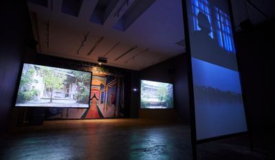 Three video installation screens showing residential buildings, a woman's shadow, and a nightscape, in front of mural 