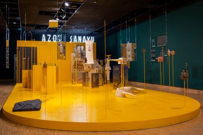 A yellow podium features a series of poles and plexiglass sheets that support totemic sculptures and other archival materials in an installation by Cooking Sections.