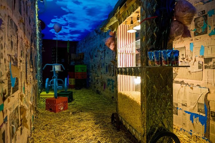 An art installation taking up a dimly lit room, with hay placed on the floor and walls either side covered in class=