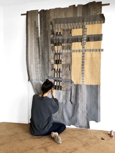 The artist Hana Miletić is pictured sitting on the ground, working on a large textile piece made up of different, woven parts.