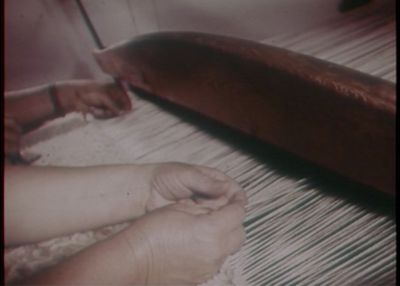 A grainy image captures the hands of women working a loom.