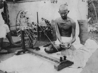 A black and white photograph captures Gandhi sitting on the floor, weaving.