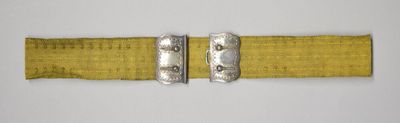 A gold silk belt with a metal buckle from the early 20th century, belonging to the Victoria & Albert Collection, is photographed against a grey background.