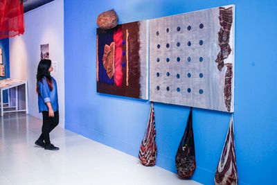 A textile work with teardrop-shaped hanging parts is placed against a blue wall, showing at the Dhaka Art Summit of 2018.