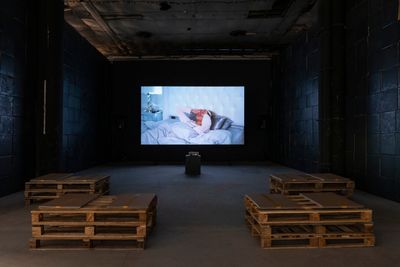 Video installation screen showing man in bed eyes covered by his hands.