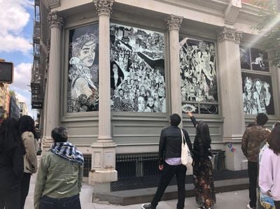 Crowd at intersection pointing at gothic black and white figure drawings printed on outer walls of building.