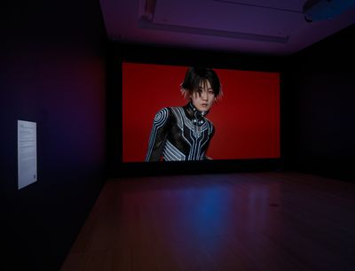 One large screen in a darkened gallery space features the artist Lu Yang dressed as her alter ego Doku in futuristic clothing, against a red background.