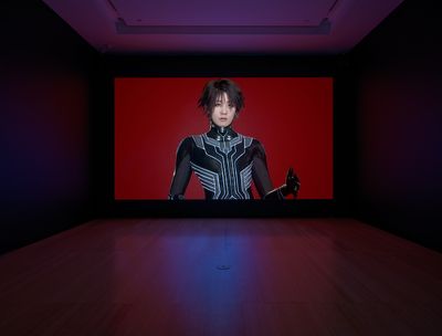 One large screen in a darkened gallery space features the artist Lu Yang dressed as her alter ego Doku in futuristic clothing, against a red background.