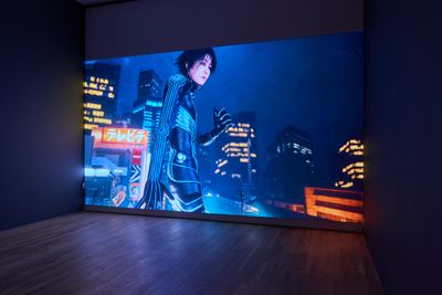 A large screen in the gallery space shows the artist Lu Yang as her alter ego, Doku, dressed in a futuristic outfit in a sci-fi landscape of screens and buildings.