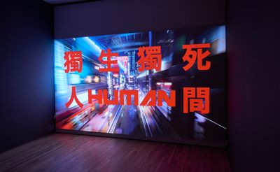 A large screen in a darkened gallery space features a futuristic scene, over which Chinese characters are written in red.