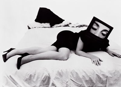 A black and white image of a heeled figure lying across a bed is captured with a television as their head. In the frame of the screen, it is just their closed eyes that are visible.