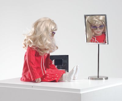 A doll with blonde hair and wearing sunglasses is dressed in a pink tracksuits and sits atop a white table top, in front of a small mirror.