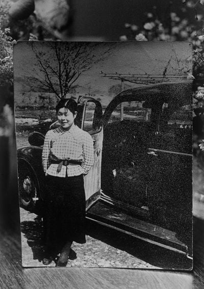 A black and white photograph of Miyako Ishiuchi's mother captures her standing in front of a vehicle, smiling at the camera.
