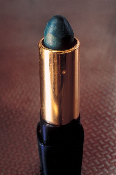A blue-grey lipstick in a golden case is photographed up close with the lid off, standing on a table.