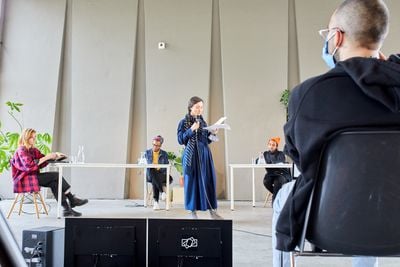 Monika Szewczyk, director of de Appel stands on a stage with a microphone surrounded by speakers sitting at tables behind her. She is opening Home is Where the Music Is, a seminar series at de Appel, conceived by the curatorial team of sonsbeek20→24.