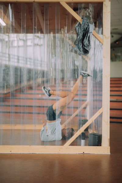 SofiCca lies on her back with her legs kicked over her head. She is performing within a wooden structure covered in plastic sheeting at de Appel.