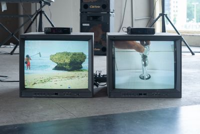 Two television units on the floor on the exhibition space feature a man standing in the ocean taking field recordings with a microphone, to the left, and a running tap to the right.
