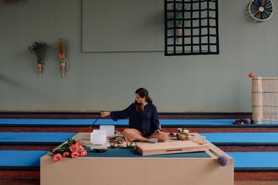 Ro Acosta sits on a platform playing a sound bowl. She is surrounded by sound bowls and a bouquet of pink flowers sits to her right, at the front of the platform.
