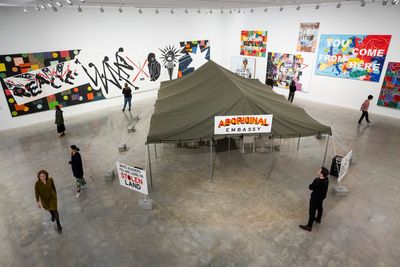 A series of paintings in the background of a gallery space is foregrounded by a tent titled 'Aboriginal Embassy',  in front of which a sign reads 'White Invaders You Are Living On Stolen Land'
