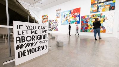 A series of paintings in the background of a gallery space is foregrounded by a sign that reads 'If you can't let me live Aboriginal, why preach democracy?'