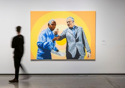 A painting by Richard Bell in a gallery space of two men in suits against an orange and yellow background.