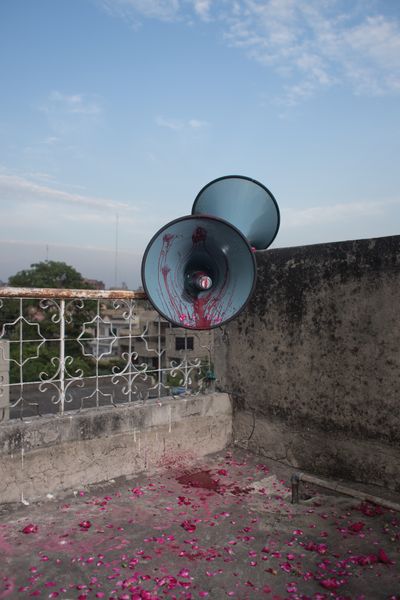 A colour photograph captures a megaphone positioned in the corner of an outdoor terrace space. Purple liquid and flowers spill from its interior onto the floor.