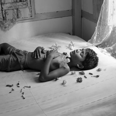 A young boy is photographed lying on a bed with hibiscus flowers strewn across him and the bed.