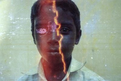 A colour photograph by Sathish Kumar captures the face of a young boy looking out towards the camera. His face is distorted with a light glare.