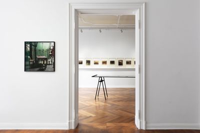 Small photograph depicting domestic interiors in front of a second exhibition space containing a row of framed prints and large table. 
