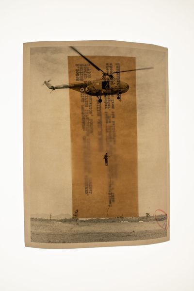 Pigment print showing a soldier descending from helicopter in front of a sheet of brown paper with text.