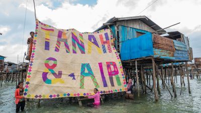 Two women stand in the water below a cluster of houses on stilts, and another woman stands on a platform above them. The three women hold up a large mat that reads 'Tanah & Air'. The writing is colourful and patterned with stripes and diamonds, and stands brightly against the beige background of the mat. There is a small dolphin between '&' and 'Air'.