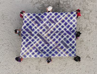 A purple and white criss-crossed mat featuring images of tables on its surface is held up by eight people standing on the sand. The supported mat is photographed from above.