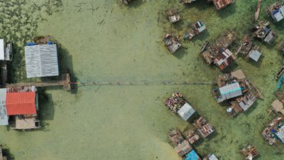 An aerial shot features the homes of a sea village, made up of houses on stilts. Two clusters of buildings are joined by a long narrow woven mat that has been unravelled by its carriers.