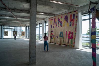 An industrial gallery space with large windows running along its lengths features an immense mat that hangs from floor to ceiling and features a small dolphin along with the words 'Tanah & Air'.