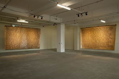 Two large mats, beige in colour, feature illegible writing across their surfaces. They are hung in a dimly lit gallery room.