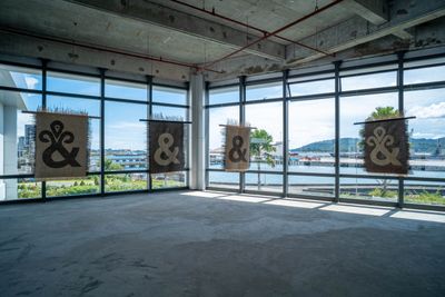 Four mats hang from the ceiling against windows looking out to sea. Each mat, created by Yee I-Lann and her collaborators, features the '&' sign, with shapes that have been reinterpreted. 