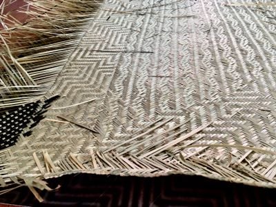 A mat in the process of being woven is photographed on the floor close up, so that the unfinished, frayed ends of the threads are visible, as is a died black patterned segment.