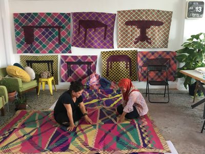 Artist Yee I-Lann and one of her collaborators are inspecting a colourful patterned mat that has been laid out on the floor in a hip, furnished room.