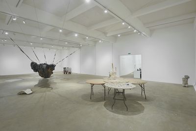 An installation by Yu Ji in the gallery space, featuring a set of three round tables with sculptures placed on top, and a black hammock-like structure made of netting and tarpaulin stretching from one side of the gallery to the other.
