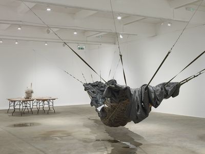 An installation by Yu Ji in the gallery space features a large black hammock-like structure hanging from wall to wall, and a set of five circular overlapping tables support a torso made from cement at their centre. The table are to the left, and the hammock is to the right.
