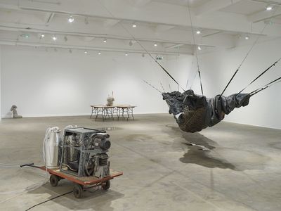 An installation by Yu Ji in the gallery space features three distinct works: a large hammock-like structure to the left, which hangs from wall to wall, a set of five circular overlapping tables with a torso made from cement at their centre, to the far left, and at the front, there is a machine made up of two milk canisters that sit atop a trolley.