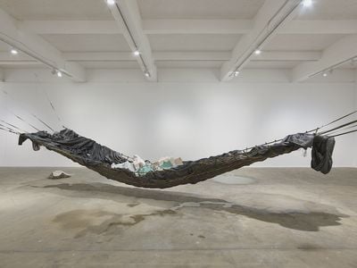 A large hammock-like structure hangs from wall to wall in the gallery space, with rubble placed in its centre and pools of water beneath it on the floor.