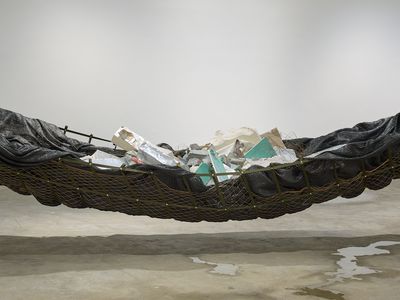 A large hammock made from netting and black tarpaulin is closely cropped, so that the rubble placed inside it is visible, as are pools of water beneath it.