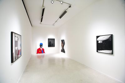 In a white gallery space, framed black and white photographs by Zanele Muholi sit along the walls. At the far end of the gallery, a bright red portrait sits against the wall on the floor.