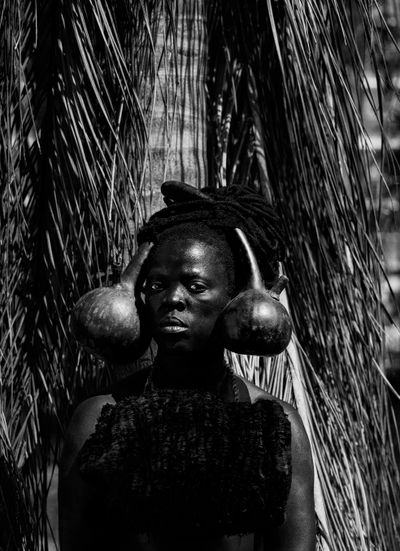A portrait of artist Zanele Muholi captures the artist with two gourds resting either side of their head. Their gaze fixes the viewer.